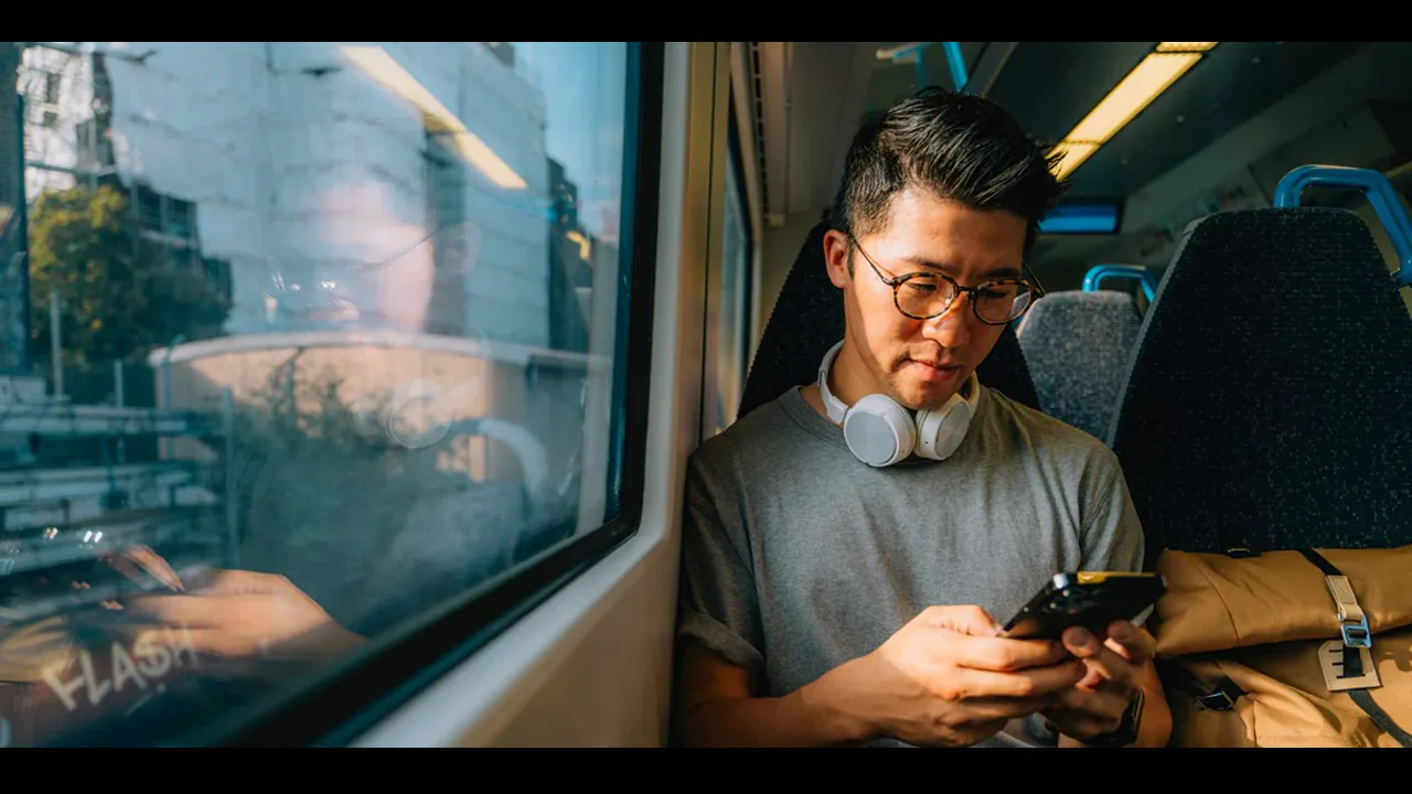 A man wearing headphones around his neck watching his phone on a train